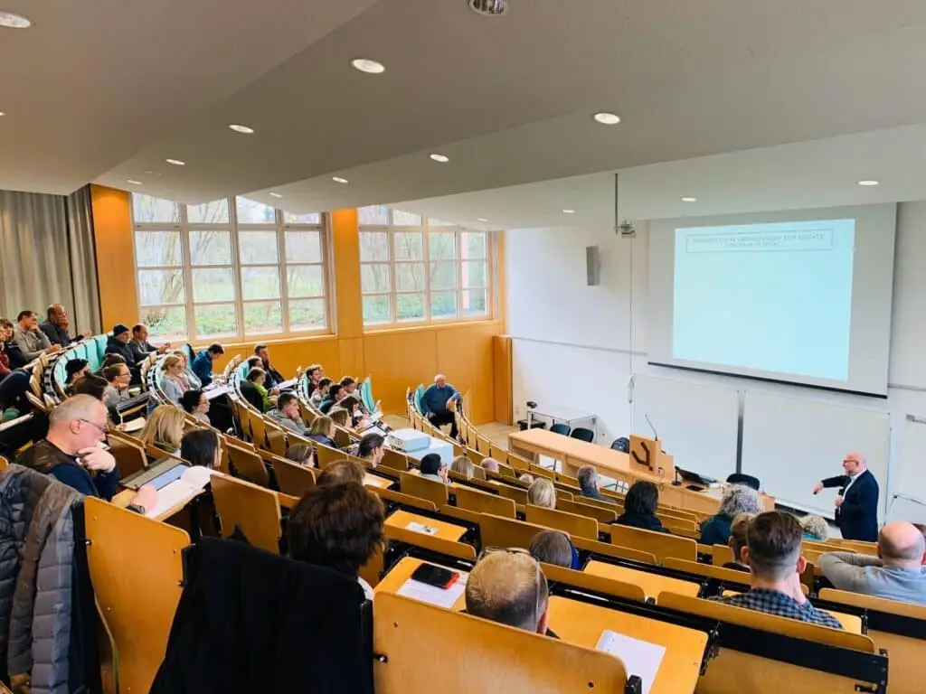 Ein Dozent hält einen Vortrag in einem gut besuchten Hörsaal. Die Zuhörer sitzen in gestaffelten Sitzreihen und machen Notizen. An der Wand ist eine Präsentation mit dem Titel „Wirtschaftliche Überlegungen zum Einsatz von Marketing“ zu sehen. Große Fenster lassen Tageslicht herein.