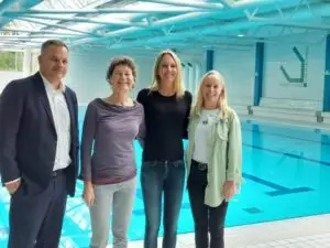 Vier Personen stehen lächelnd vor einem Hallenbad mit blauem Wasser. Im Hintergrund ist eine Sitztribüne und die Aufschrift „Sportschule“ zu sehen. Die Gruppe umfasst zwei Männer und zwei Frauen in Freizeit- und Geschäftskleidung.