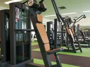 Mehrere moderne Fitnessgeräte in einem hellen, geräumigen Trainingsraum mit grünem und schwarzem Boden. Polsterungen der Geräte sind beige. Spiegel an der Wand reflektieren den Raum, der gut beleuchtet ist.