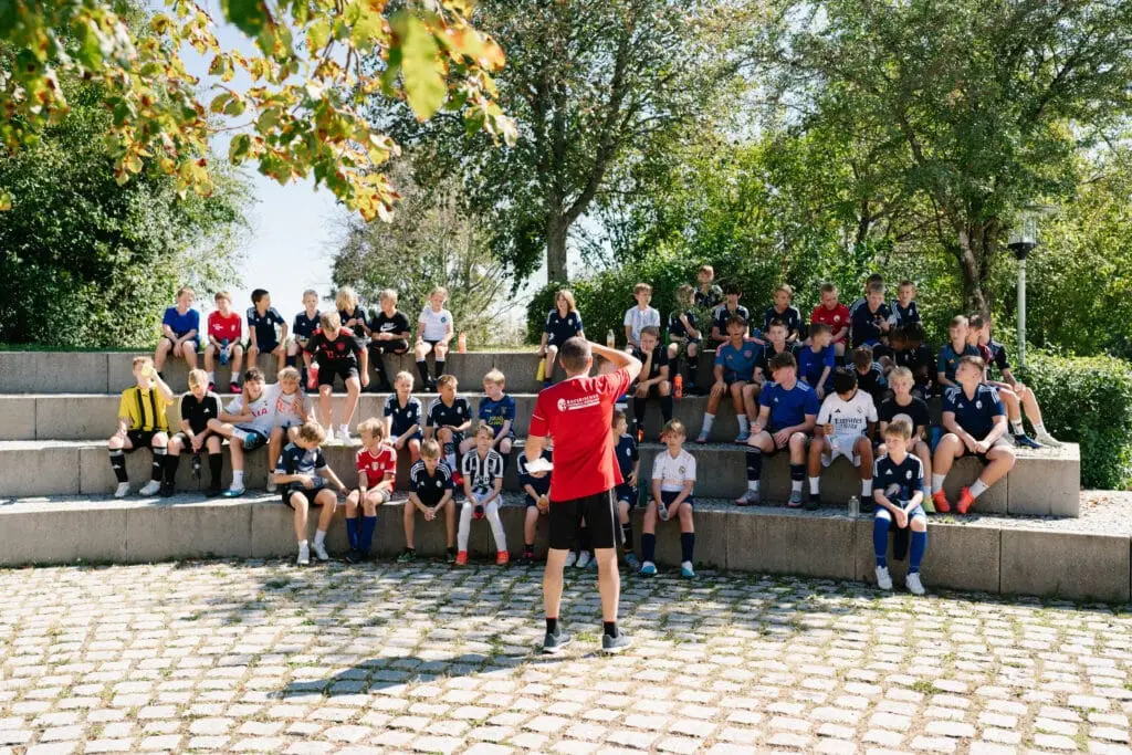 Junge Fußballspieler sitzen auf Stufen einer gepflasterten Tribüne im Freien. Ein Trainer in einem roten Shirt steht vorne und spricht zu ihnen. Die Szene ist von Bäumen eingefasst, die für Schatten sorgen.