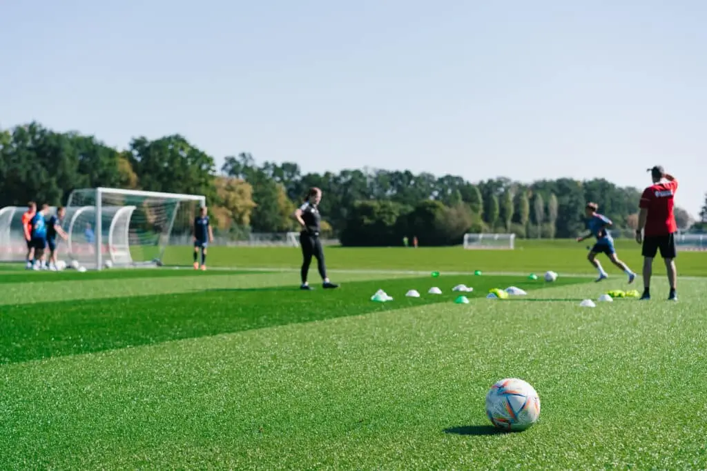 Auf einem Fußballplatz trainieren mehrere Personen, umgeben von grünen und weißen Hütchen. Im Hintergrund stehen Tore und Bänke, während ein Fußball im Vordergrund liegt.
