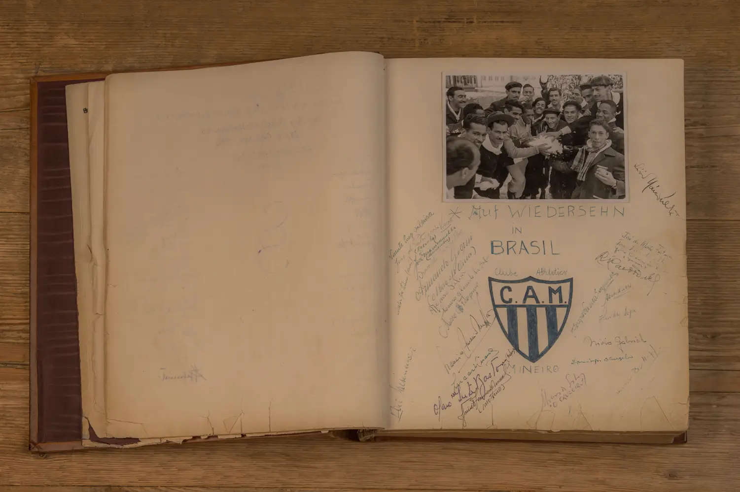 Offenes Buch mit einer Schwarz-Weiß-Fotografie von Menschen, die sich umarmen. Darunter steht "Auf Wiedersehen in Brasil." Es gibt mehrere Unterschriften in verschiedenen Farben und ein Wappen mit der Aufschrift "C.A.M. Clube Atlético Mineiro.