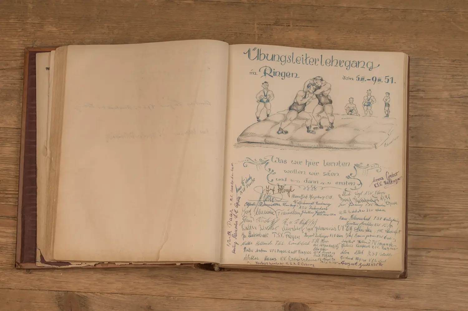 Geöffnetes Buch auf einem Holztisch zeigt eine Seite mit einer handgezeichneten Illustration von Ringern und Beschriftungen in deutscher Schrift. Über der Illustration steht "Übungsleiterlehrgang im Ringen", darunter das Datum "vom 5.-9. III. 51.". Darunter befindet sich der Text "Was wir hier lernten, wollen wir säen und dann ernten", umgeben von zahlreichen Unterschriften.