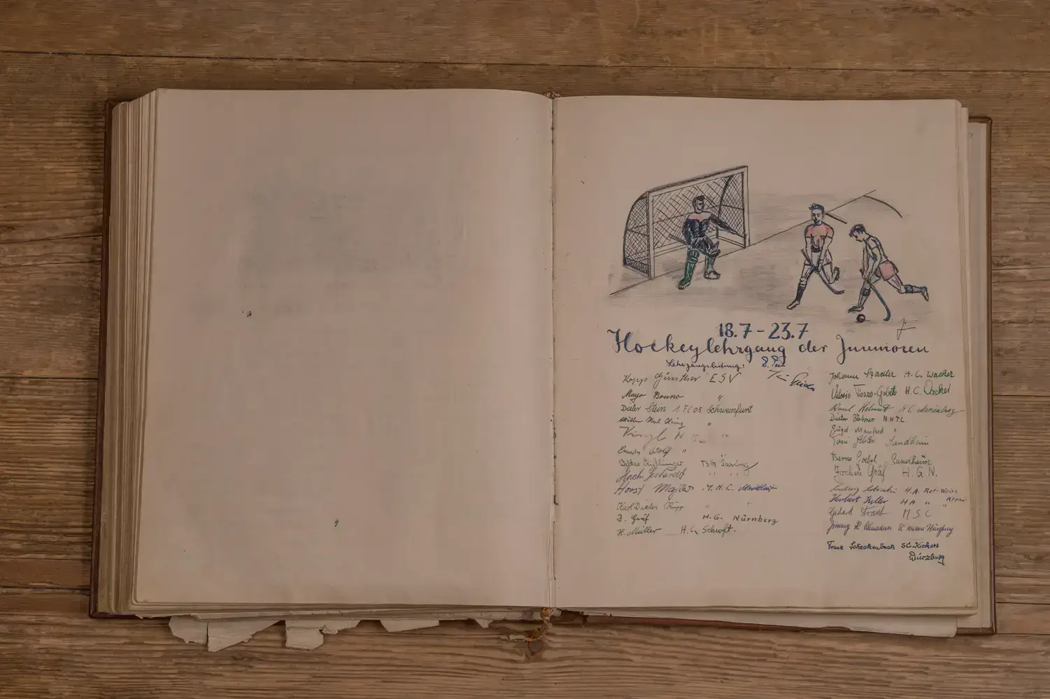 Offenes Buch auf einem Holztisch. Rechte Seite zeigt eine handgezeichnete Illustration eines Hockeyspiels mit drei Spielern vor einem Tor. Darunter handschriftlicher Text und Datum 18.7 - 23.7 mit mehreren Namen und Unterschriften in verschiedenen Farben. Linke Seite leer bis auf einige verblasste Textspuren.