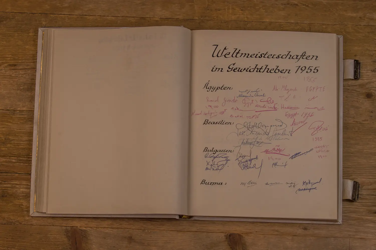 Offenes Buch auf Holztisch mit der Überschrift „Weltmeisterschaften im Gewichtheben 1955“ auf der rechten Seite. Darunter handschriftliche Einträge und Unterschriften von Teilnehmern aus Ländern wie Ägypten, Brasilien, Bulgarien und Burma.