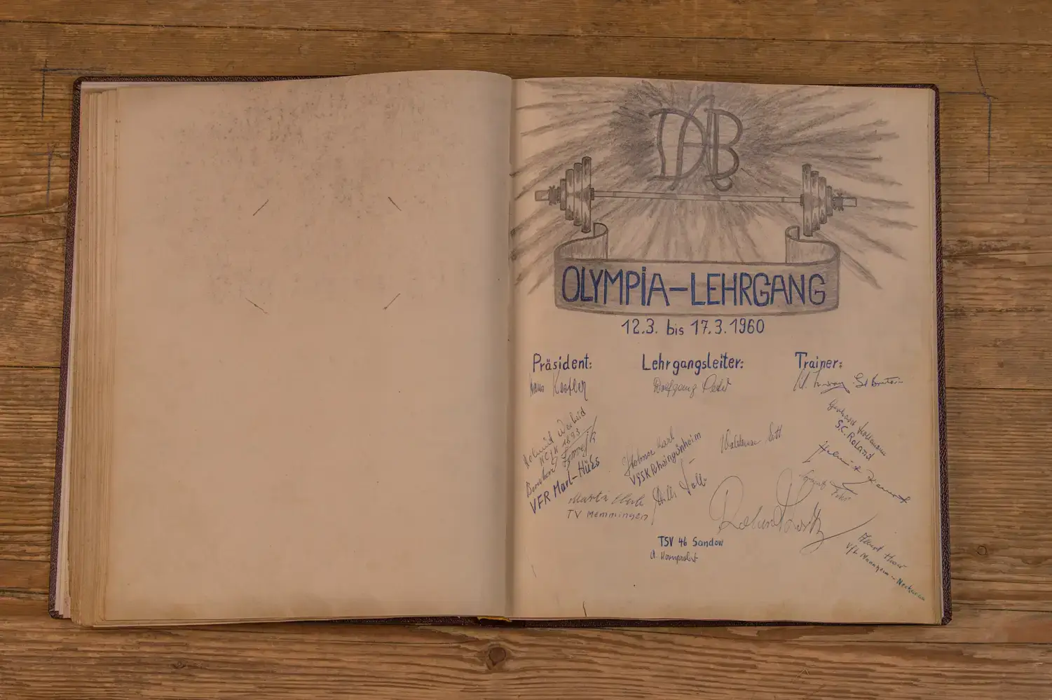 Offenes Buch auf Holztisch. Rechte Seite zeigt eine handgezeichnete Illustration mit einer Hantel und den Worten "Olympia-Lehrgang 12.3. bis 17.3.1960". Darunter sind die Titel "Präsident", "Lehrgangsleiter" und "Trainer" mit handschriftlichen Namen und Unterschriften versehen. Links ist eine leere Seite.