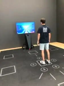 Ein junger Mann steht auf einem interaktiven Bodenmatten-Spielsystem mit geometrischen Markierungen, während er auf einen Bildschirm vor sich blickt, auf dem ein digitales Laufspiel gezeigt wird. Der Raum ist minimalistisch eingerichtet.