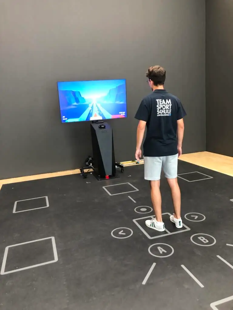 Ein junger Mann steht auf einem interaktiven Bodenmatten-Spielsystem mit geometrischen Markierungen, während er auf einen Bildschirm vor sich blickt, auf dem ein digitales Laufspiel gezeigt wird. Der Raum ist minimalistisch eingerichtet.