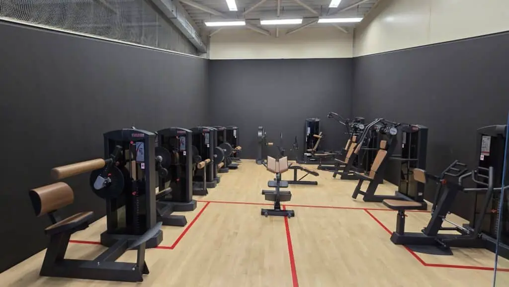 Raum mit mehreren Fitnessgeräten, die an den Wänden aufgestellt sind. Der Boden ist aus Holz, und die Geräte haben überwiegend schwarze und braune Polster. Deckenbeleuchtung und ein Netz an der oberen Wand.