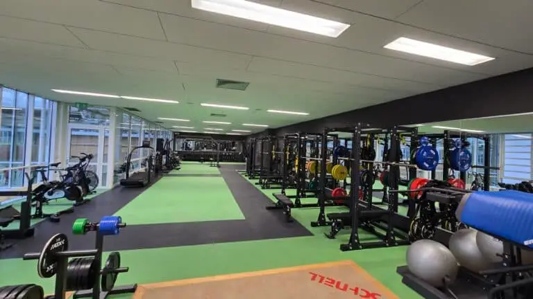 Großer, gut ausgestatteter Fitnessraum mit mehreren Gewichtsständern, Hantelbänken und Gymnastikbällen. Links sind stationäre Fahrräder und Laufbänder zu sehen. Rechts befinden sich Schwerlastregale mit bunten Gewichtsscheiben. Der Boden ist mit grünen und schwarzen Matten belegt. Fensterfronten bieten viel Tageslicht.
