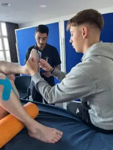 Eine Person sitzt auf einer Liege und misst mit einem Messwerkzeug das Bein eines anderen, auf dem blaues Kinesiologie-Tape angebracht ist. Eine dritte Person steht daneben und beobachtet. Im Hintergrund ein Raum mit großem Fenster und blauen Trennwänden.