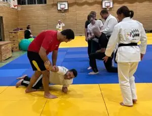 Mehrere Menschen befinden sich in einer Sporthalle mit Judomatten. Zwei Männer üben eine Verteidigungstechnik; einer kniet nieder und hält den Arm des anderen, der auf der Matte liegt. Andere Personen stehen um sie, einige in Judogi, und scheinen zuzuschauen oder Anweisungen zu geben. Im Hintergrund sind Turnkästen, Sitzbälle und Basketballkörbe zu sehen.