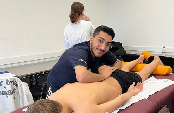 Ein Mann führt an einem liegenden Patienten auf einer Massagebank eine manuelle Therapie durch. Im Hintergrund sitzt eine weitere Person auf einem Stuhl.