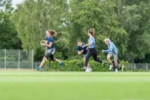 Mehrere Personen spielen Frisbee auf einem grünen Feld. Eine Person hält die Frisbee, während drei andere hinter ihr herlaufen. Im Hintergrund sind Bäume und ein Zaun zu sehen.