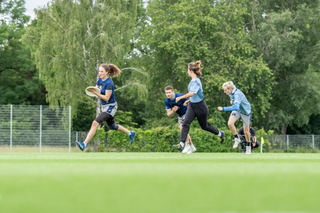 Eine Gruppe von Personen spielt Frisbee auf einem grünen Spielfeld, umgeben von Bäumen. Eine Person in der Mitte läuft mit der Frisbee in der Hand, während andere spielerisch versuchen, sie einzuholen. Die Szene zeigt sportliche Aktivität und Bewegung.