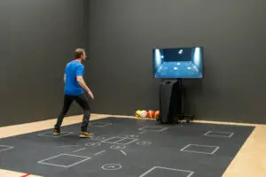 Ein Mann in einem blauen T-Shirt steht auf einer Bodenmatte mit markierten Quadraten und schaut auf einen Monitor, der ein Spiel mit Bällen darstellt. Neben dem Monitor liegen verschiedene Sportbälle auf dem Boden.