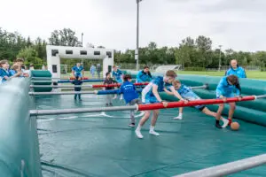 Menschen spielen gemeinsam auf einem gigantischen Tischkickerfeld im Freien. Sie bewegen sich entlang von Stangen, ähnlich wie bei einem Tischfußballspiel. Die Spieler tragen sportliche Kleidung und zeigen Freude beim Spielen. Im Hintergrund ist ein aufblasbares Fußballtor und eine grüne Sportwiese zu sehen.