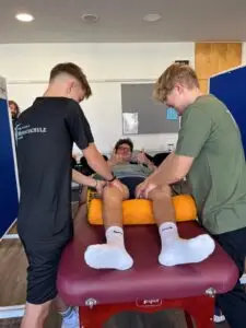 Ein Mann liegt lächelnd auf einem Massagetisch, während zwei junge Therapeuten seine Beine massieren. Der Mann zeigt zwei Daumen hoch und scheint zufrieden. Im Hintergrund ist ein Raum mit blauen Trennwänden und hellen Wänden.