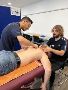Zwei Männer behandeln eine Person, die mit freiem Oberkörper auf einem Massagetisch liegt. Einer drückt auf den Rücken, während der andere den Kopf der liegenden Person hält. Im Raum stehen ein blauer Paravent mit der Aufschrift "TEAM B", einige Stühle und Handtücher.