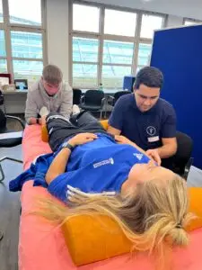 Eine Frau liegt auf einer Liege und wird von zwei jungen Männern untersucht. Die Frau trägt ein blaues Trikot mit einem Logo, während die Männer sportliche Kleidung tragen. Der Raum hat große Fenster im Hintergrund.