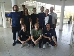 Eine Gruppe von zehn Menschen, gekleidet in sportliche T-Shirts mit einem Logo, steht und kniet in einem modernen, lichtdurchfluteten Gebäude. Sie lächeln in die Kamera. Im Hintergrund sind Glaswände und ein Treppenhaus sichtbar.