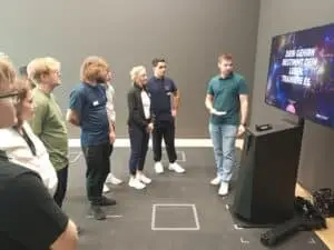 Eine Gruppe von Menschen steht in einem Raum vor einem großen Bildschirm, der die Worte "Dein Gehirn bestimmt dein Leben. Trainiere es." zeigt. Ein Mann hält eine Präsentation. Die Anwesenden hören aufmerksam zu.