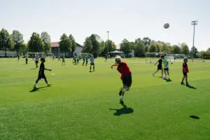 Kinder spielen Fußball auf einem weitläufigen, sonnigen Spielfeld. Einige Kinder tragen Trainingsleibchen, und ein Kind wirft den Ball ein. Im Hintergrund sind Tore und Bäume zu sehen, die das Spielfeld umgeben.