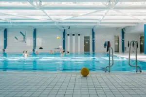 Ein Hallenbad mit mehreren Personen, die im Wasser Polo spielen. Ein gelber Wasserball liegt am Beckenrand vor einer Einstiegsleiter. Im Hintergrund sind Umkleidekabinen und Handtücher sichtbar. Die Decke ist mit weißen Stahlträgern versehen, durch die Tageslicht einfällt.