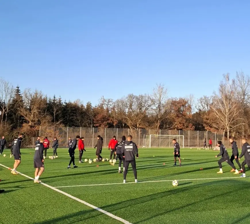 Gruppe von Fußballspielern beim Training auf einem Rasenplatz, umgeben von Bäumen. Die Spieler tragen Sportbekleidung in Rot und Schwarz und üben mit mehreren Fußbällen bei sonnigem Wetter. Im Hintergrund ist ein Tor zu sehen.