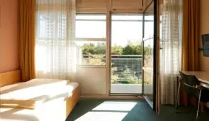 Ein sonnendurchflutetes Zimmer mit einem Einzelbett links, einem großen Fenster mit geöffneter Tür in der Mitte, die zu einem Balkon führt, und einem Schreibtisch mit Stuhl rechts. Gelbe Vorhänge und eine grüne Pflanzenlandschaft sind draußen sichtbar.