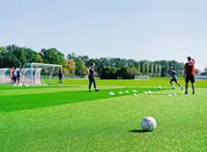 Mehrere Personen trainieren auf einem Fußballfeld. Ein Ball liegt im Vordergrund, während im Hintergrund Spieler Übungen absolvieren und ein Trainer Anleitung gibt. Im Hintergrund sind Tore und grüne Bäume zu sehen.