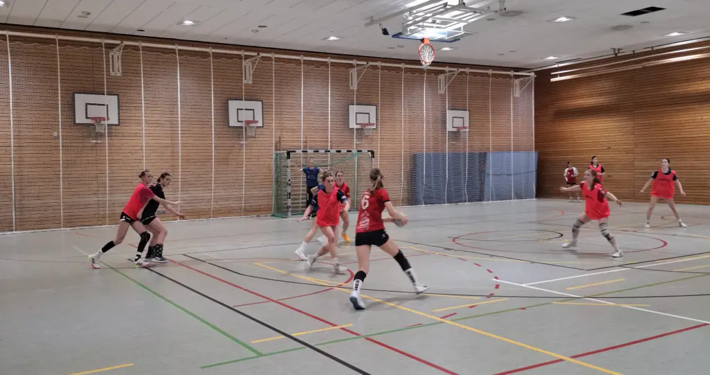 Frauen-Handballspiel in einer Sporthalle, sechs Spielerinnen in rot-schwarzen Trikots mit Ball in Richtung Tor. Gegnerische Mannschaft in blau versucht zu verteidigen. Spielerinnen sind in Bewegung, Sporthalle mit Holzwand und mehreren Basketballkörben im Hintergrund.