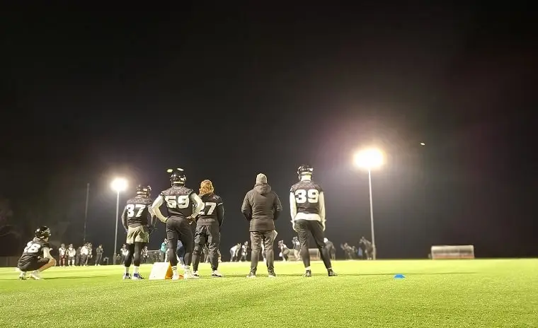 Mehrere American-Football-Spieler in voller Ausrüstung stehen auf einem beleuchteten Spielfeld bei Nacht. Ein Trainer, in der Mitte, hält eine Ansprache an die Spieler. Im Hintergrund sind weitere Personen und große Flutlichter sichtbar.