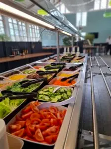 Eine Salatbar mit verschiedenen Schalen gefüllt mit frischen Zutaten wie Tomaten, Gurken, Karotten, Salatblättern und Mais. Im Hintergrund ist ein helles, modernes Essbereich mit großen Fenstern und Pflanzen zu sehen.