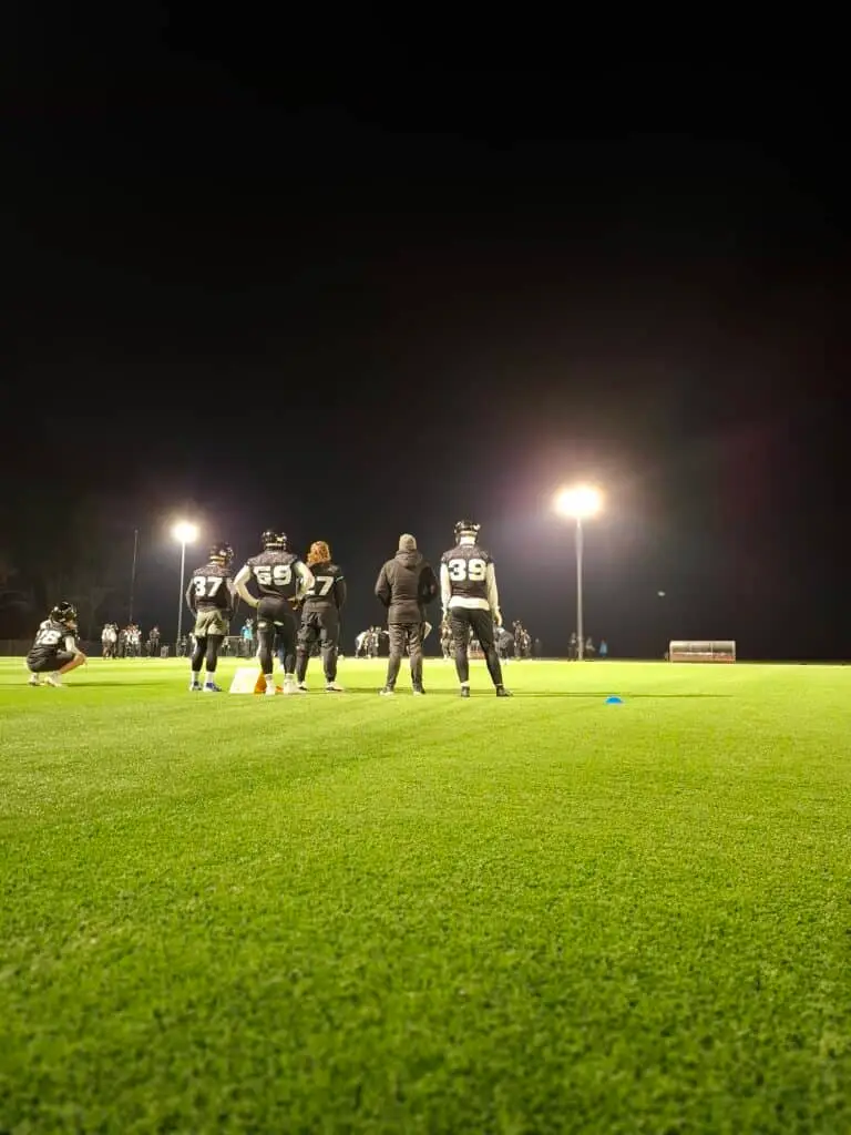 Mehrere American-Football-Spieler und ein Trainer stehen auf einem beleuchteten Sportfeld, umgeben von Flutlichtern in der Nacht. Sie sind in Ausrüstung gekleidet und scheinen an einer Trainingsübung teilzunehmen, während im Hintergrund weitere Spieler und Personen zu sehen sind.