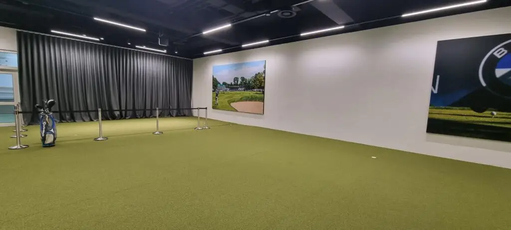 Innenraum mit grünem Teppichboden, einigen Absperrungen und einem Golfschläger-Set links. An der Wand hängen zwei große Bilder, eines zeigt eine Golfplatzlandschaft. Im Hintergrund schwarze Vorhänge.