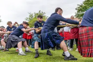 Mehrere Personen in Kilts ziehen in einem Tauziehen mit, wobei eine Gruppe Zuschauer im Hintergrund zu sehen ist. Die Anstrengung der Teilnehmer ist erkennbar, während sie das Seil energisch ziehen. Die Umgebung ist grün und es scheint eine Freizeitveranstaltung im Freien zu sein.