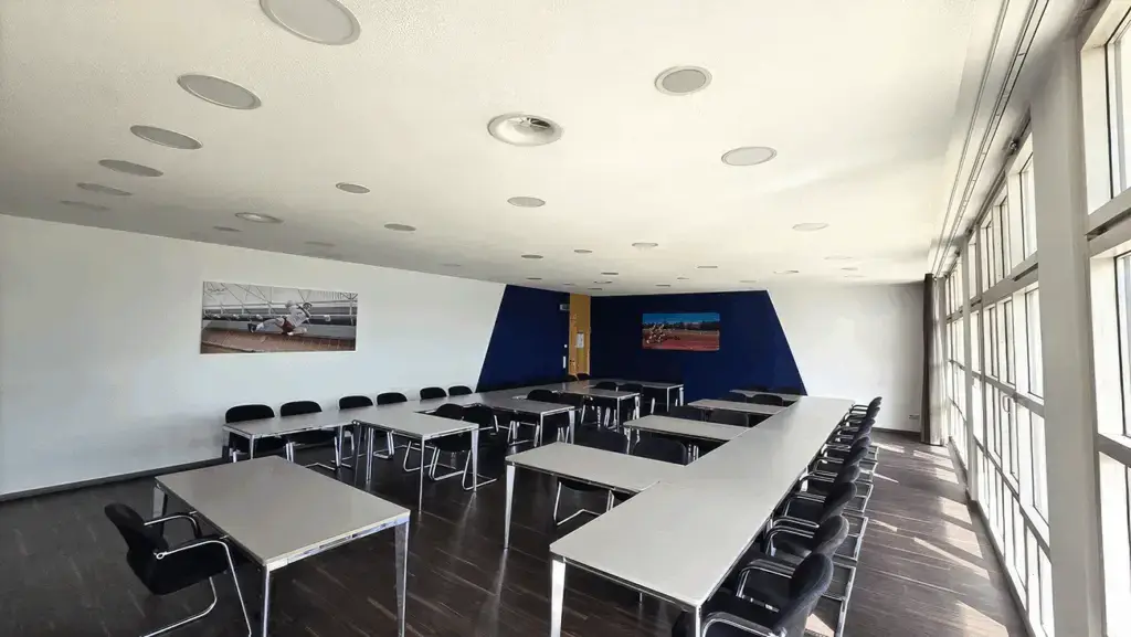 Konferenzraum mit U-förmig angeordneten Tischen und schwarzen Stühlen. Große Fenster auf der rechten Seite lassen viel Licht herein. An den Wänden hängen zwei Fotos mit Sportmotiven. Decke mit eingebauten Lautsprechern und Belüftungselementen.