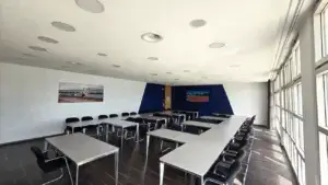 Konferenzraum mit U-förmig angeordneten Tischen und schwarzen Stühlen. Große Fenster auf der rechten Seite lassen viel Licht herein. An den Wänden hängen zwei Fotos mit Sportmotiven. Decke mit eingebauten Lautsprechern und Belüftungselementen.
