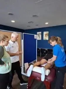 Drei Personen in einem Raum, zwei stehen links und beobachten eine Frau in blauer Arbeitskleidung, die eine sportliche Massage an den Beinen einer liegenden Person durchführt. Der Raum hat blaue Wände und Sportposter.