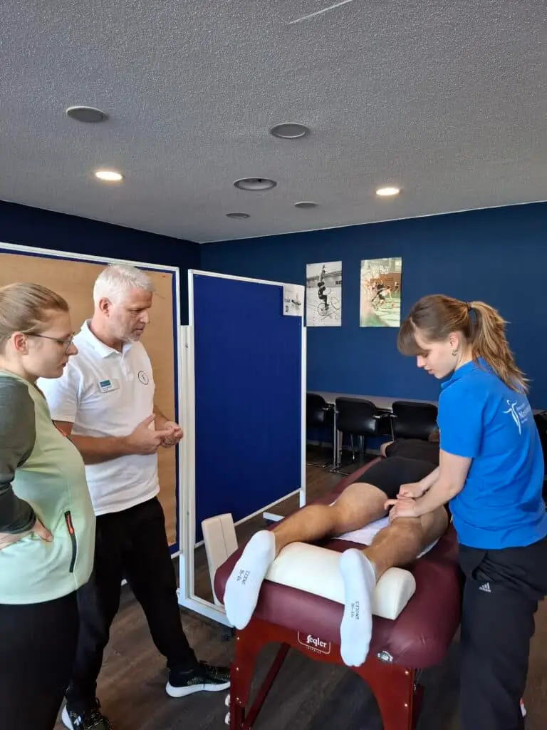 Drei Personen in einem Raum, zwei stehen links und beobachten eine Frau in blauer Arbeitskleidung, die eine sportliche Massage an den Beinen einer liegenden Person durchführt. Der Raum hat blaue Wände und Sportposter.