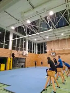 Turnhalle mit blauen Matten auf dem Boden. Eine Person führt einen Salto aus, während mehrere andere Personen auf Kastenpferden sitzen und zuschauen. Im Hintergrund sind Kletterringe und Basketballkörbe zu sehen.