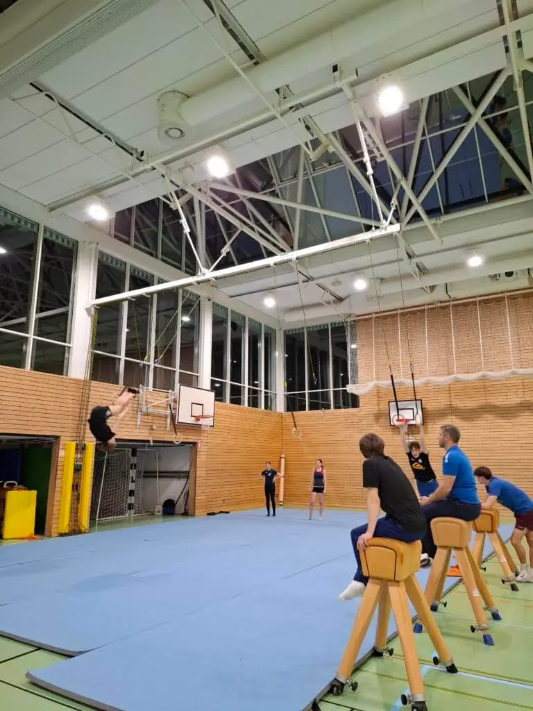 Turnhalle mit blauen Matten auf dem Boden. Eine Person führt einen Salto aus, während mehrere andere Personen auf Kastenpferden sitzen und zuschauen. Im Hintergrund sind Kletterringe und Basketballkörbe zu sehen.