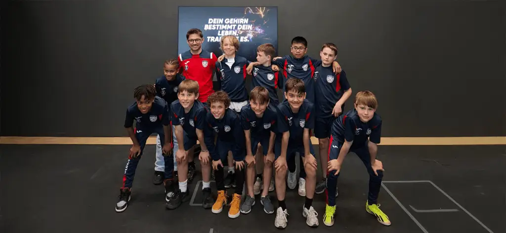 Eine Gruppe von Jungen in blauen Sporttrikots posiert mit einem Mann in einem roten Shirt vor einem Bildschirm, auf dem "Dein Gehirn bestimmt dein Leben, trainiere es" steht. Die Jungen lächeln in die Kamera, während sie sich für ein Gruppenfoto aufstellen.