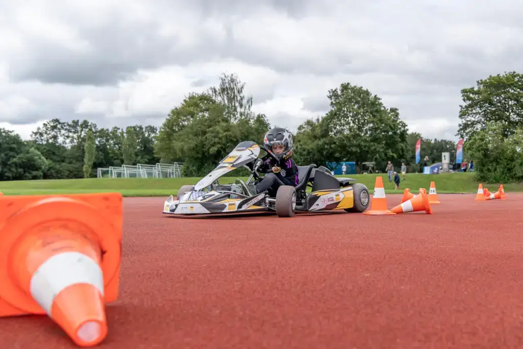 Ein Kind im Go-Kart auf einer roten Rennstrecke mit umgest&uuml;rzten Pylonen und B&auml;umen im Hintergrund