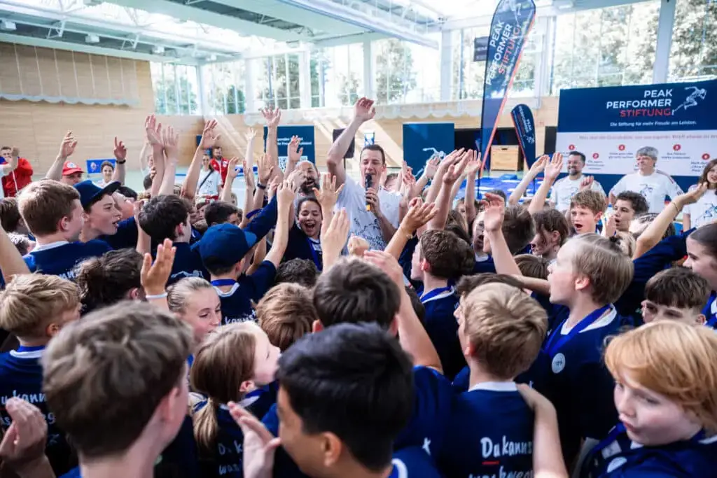 Eine große Gruppe von Kindern in blauen Trikots hebt enthusiastisch die Arme in einer Sporthalle. Im Mittelpunkt steht ein Mann mit Mikrofon, der die Kinder anfeuert. Im Hintergrund sind Banner und Erwachsene zu sehen. Die Stimmung ist lebhaft und freudig.