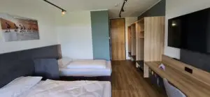 Hotelzimmer mit zwei Einzelbetten, einem großen Flachbildfernseher an der Wand, einem Schreibtisch mit Stuhl und einem offenen Kleiderschrank. Holzfußboden und teilweise grüne Wände. An einer Wand hängt ein Bild mit Personen, die auf Rollschuhen in einer Halle fahren.