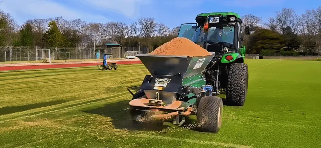 Ein grüner Traktor zieht einen Sandstreuer über ein Sportfeld, um die Rasenfläche zu pflegen. Im Hintergrund sind Bäume und eine Laufbahn sichtbar.