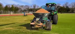 Ein grüner Traktor zieht einen Sandstreuer über ein Sportfeld, um die Rasenfläche zu pflegen. Im Hintergrund sind Bäume und eine Laufbahn sichtbar.
