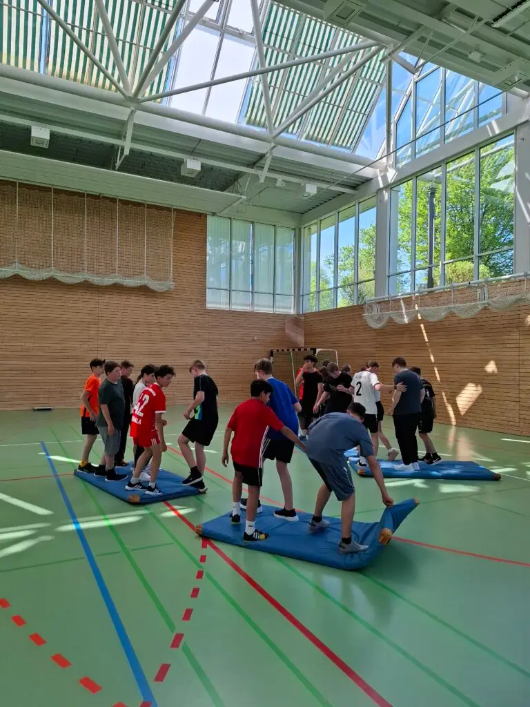 Gruppe von Jugendlichen in Sportkleidung balanciert auf Turnmatten in einer Turnhalle mit hoher Decke und großen Fenstern.